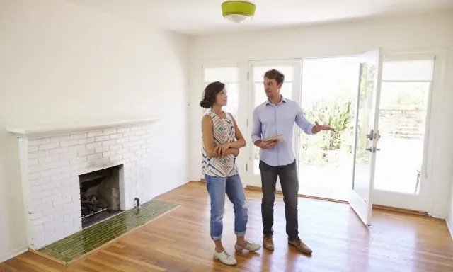 Un agent immobilier présente un appartement ou une maison à une cliente potentielle. La pièce est lumineuse, avec une cheminée en briques blanches et un sol en bois.