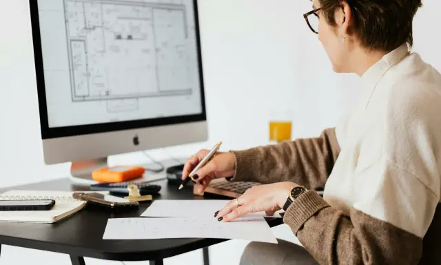 Une femme examine des plans sur son ordinateur, stylo à la main, calculatrice et notes à proximité. Elle est soulagée, car j'ai fini de payer mon crédit immobilier.