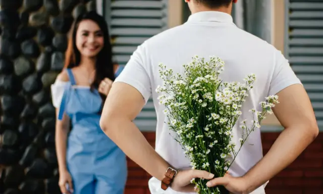 Un homme cache un bouquet de fleurs derrière son dos, prêt à surprendre sa compagne. Un couple non marié dans un lieu extérieur.