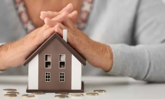 Simulation prêt immobilier après 60 ans : mains protégeant une maison miniature, avec des pièces sur la table.