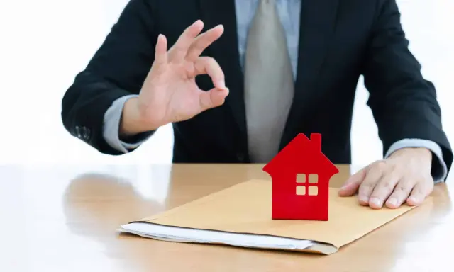 Un homme en costume fait un signe "OK" près d'une petite maison rouge sur une enveloppe. Un bon signe pour votre guide crédit immobilier.