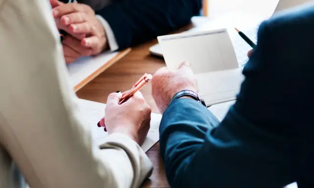 Trois mains se penchent sur un bureau en bois, stylo à la main, pour signer des documents. Le temps presse, il reste à vivre.