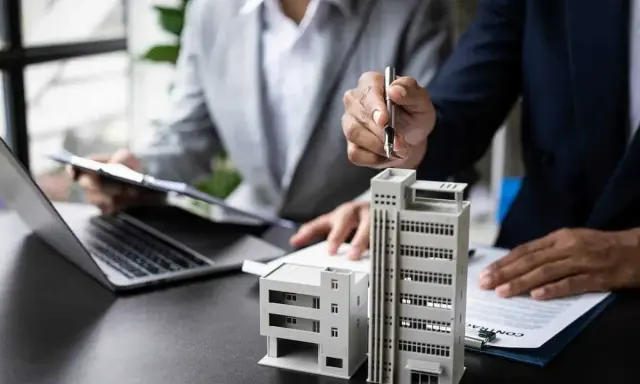 Deux personnes discutent de modèles de bâtiments et de documents, possiblement pour un projet immobilier ou un régime fiscal immobilier.