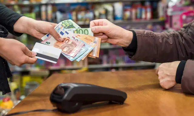 Une main tend des billets en euros et une carte bancaire. La question est : peut-on payer un artisan en liquide ?