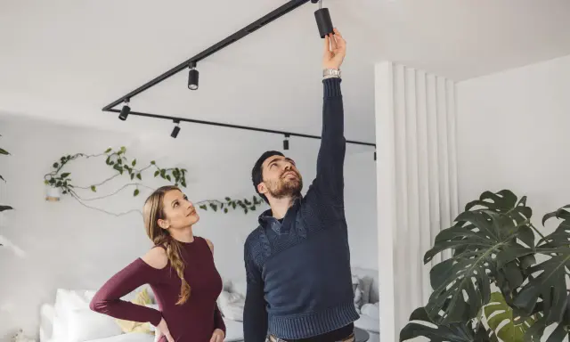 Un homme ajuste un spot sur un rail noir, sous une belle hauteur sous plafond. Une femme le regarde, curieuse.