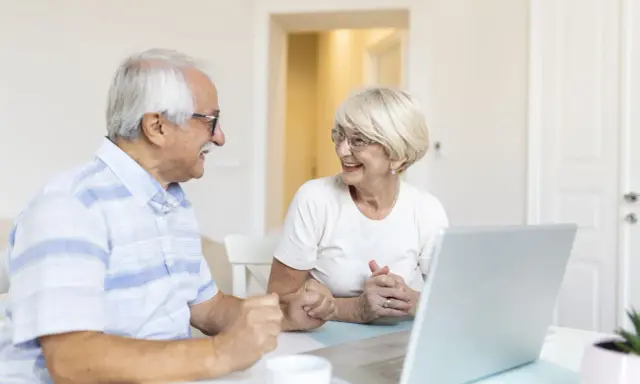 Couple âgé souriant devant un ordinateur, discutant peut-être de l'exonération taxe foncière 75 ans.