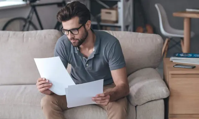 Homme concentré sur des documents, peut-être pour un crédit immobilier interimaire, assis sur un canapé.