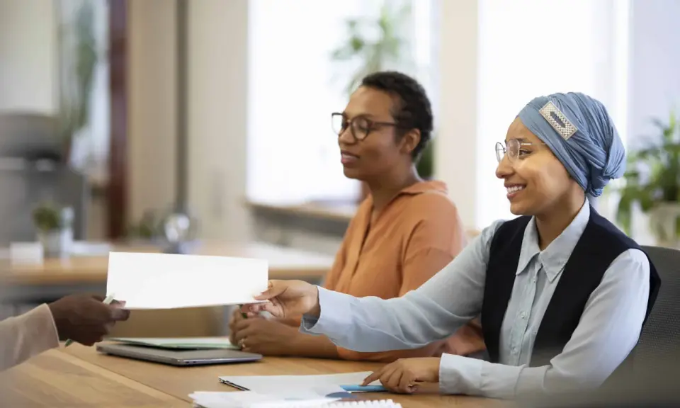 Une main tend un document blanc pour une vente en copropriété. Des femmes souriantes sont assises à une table, prêtes à examiner les documents obligatoires.