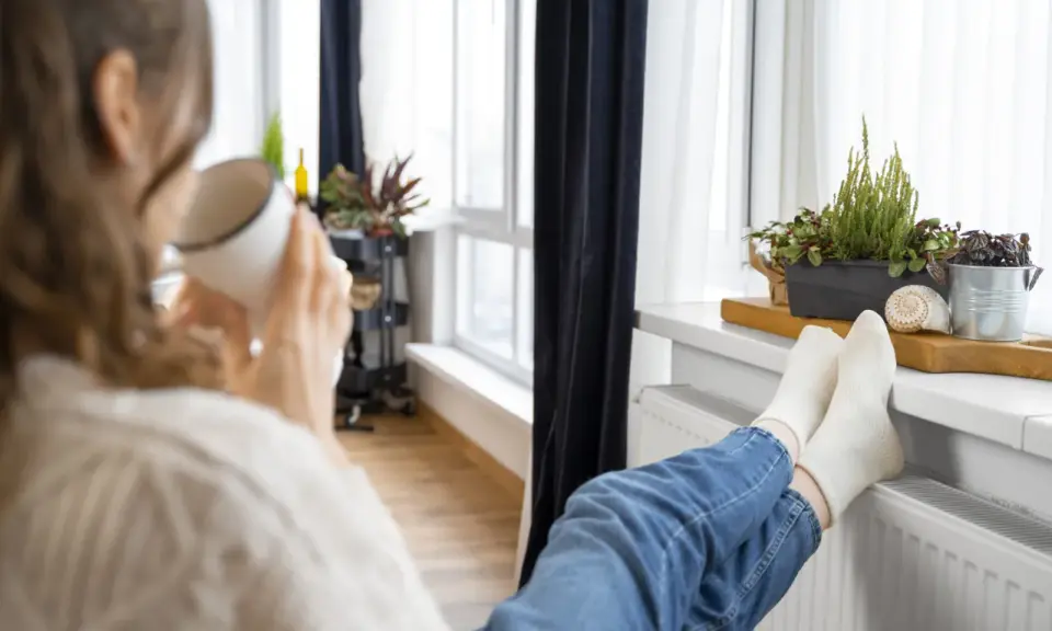 Une personne boit une tasse chaude, les pieds sur un radiateur, profitant de la chaleur. Un bon DPE chauffage électrique pour un confort optimal.