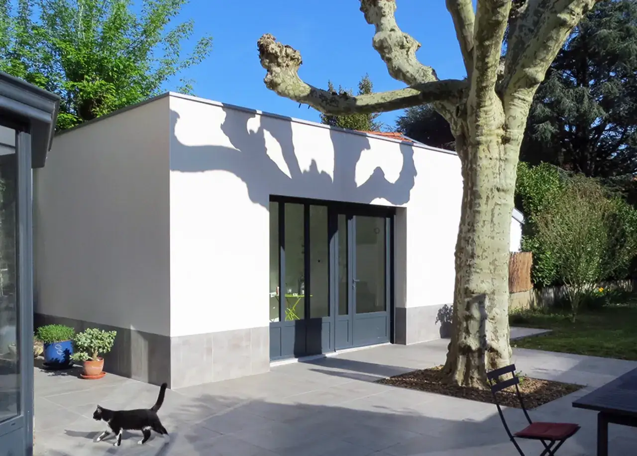 Une d&eacute;pendance moderne &agrave; c&ocirc;t&eacute; d'une maison, avec un grand platane et un chat noir. Les imp&ocirc;ts sur cette nouvelle construction sont &agrave; pr&eacute;voir.