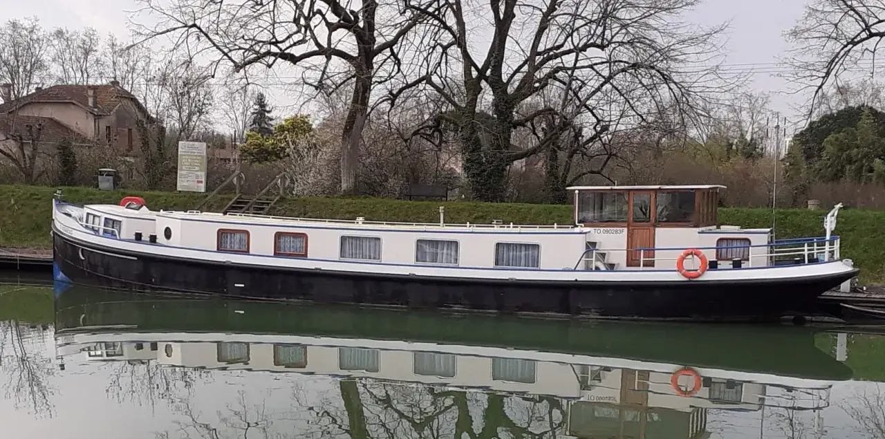 Une péniche blanche et noire, reflet d'une vie paisible sur l'eau, est amarrée près d'une maison ancienne.