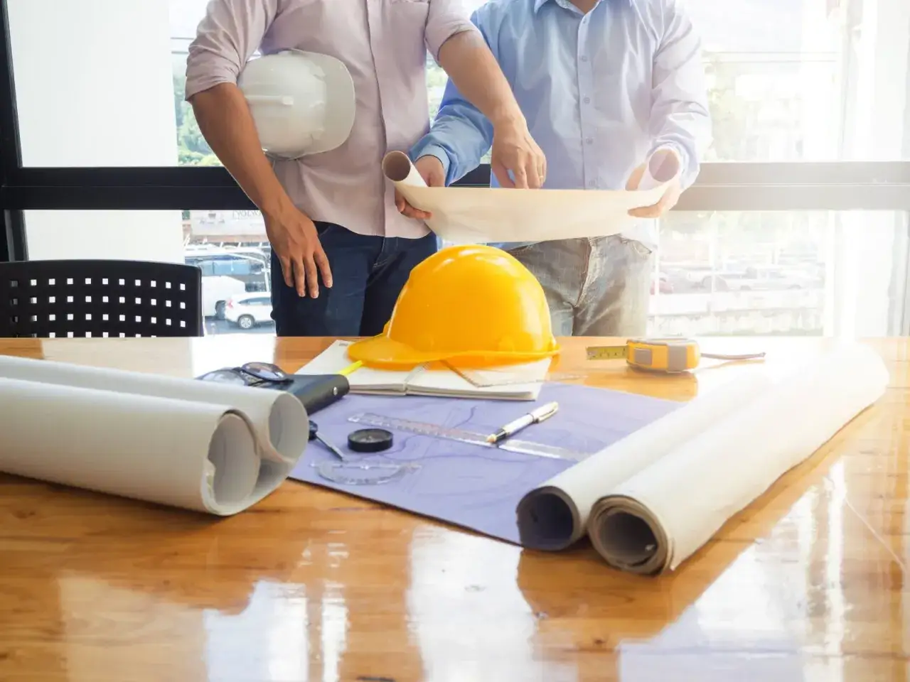 Deux hommes, un ma&icirc;tre d'&oelig;uvre et son associ&eacute;, examinent des plans sur une table de chantier. Casques, rouleaux de plans et outils sont dispos&eacute;s.