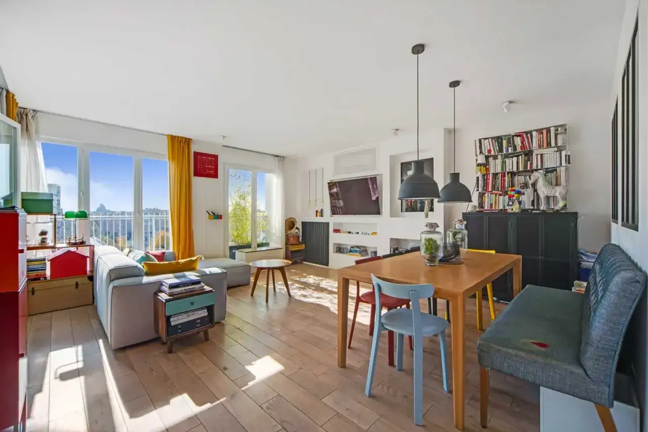 Salon lumineux d'un appartement ou maison, avec canap&eacute;, table &agrave; manger, &eacute;tag&egrave;res remplies de livres et vue sur la ville par de grandes fen&ecirc;tres.