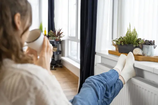 Une personne boit une tasse chaude, les pieds sur un radiateur, profitant de la chaleur. Un bon DPE chauffage électrique pour un confort optimal.