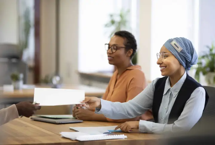 Une main tend un document blanc pour une vente en copropriété. Des femmes souriantes sont assises à une table, prêtes à examiner les documents obligatoires.