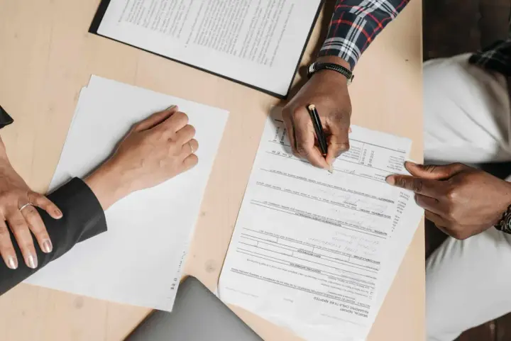 Des mains signent un compromis de vente sur un bureau en bois, entourées de documents.