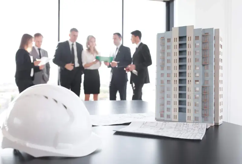 Maquette d'immeuble, plans et casque blanc sur une table. Des professionnels discutent, peut-être des travaux sur parties communes sans autorisation.