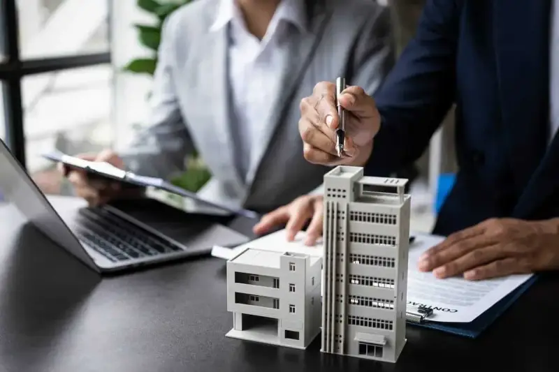 Deux personnes discutent de modèles de bâtiments et de documents, possiblement pour un projet immobilier ou un régime fiscal immobilier.